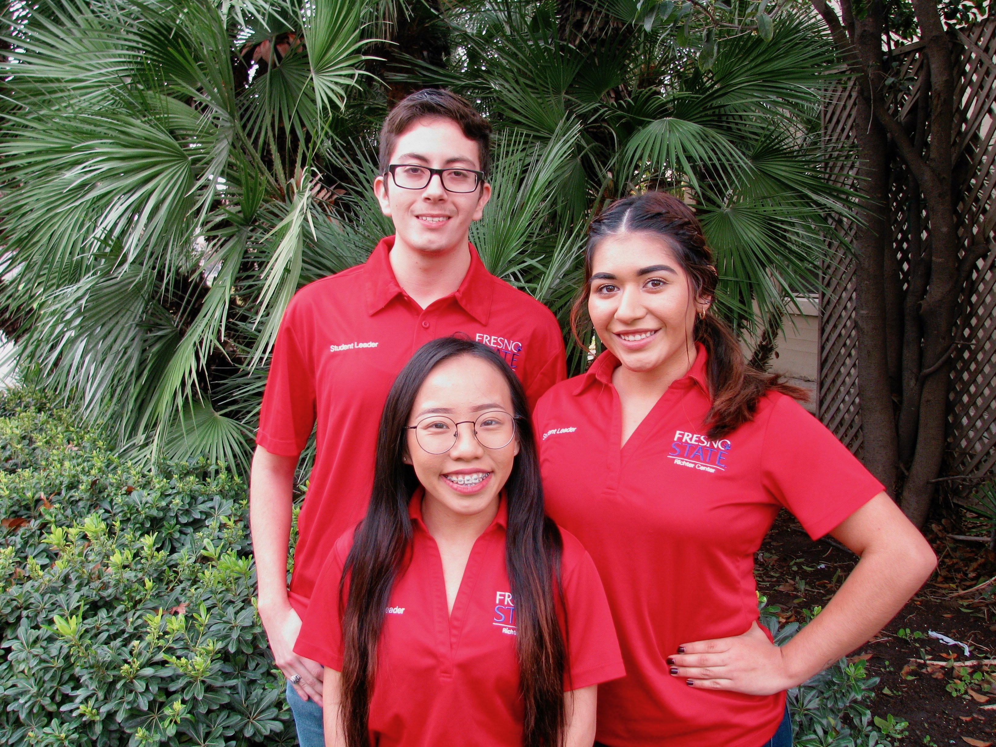 Three students stand together in front of a background with plants.