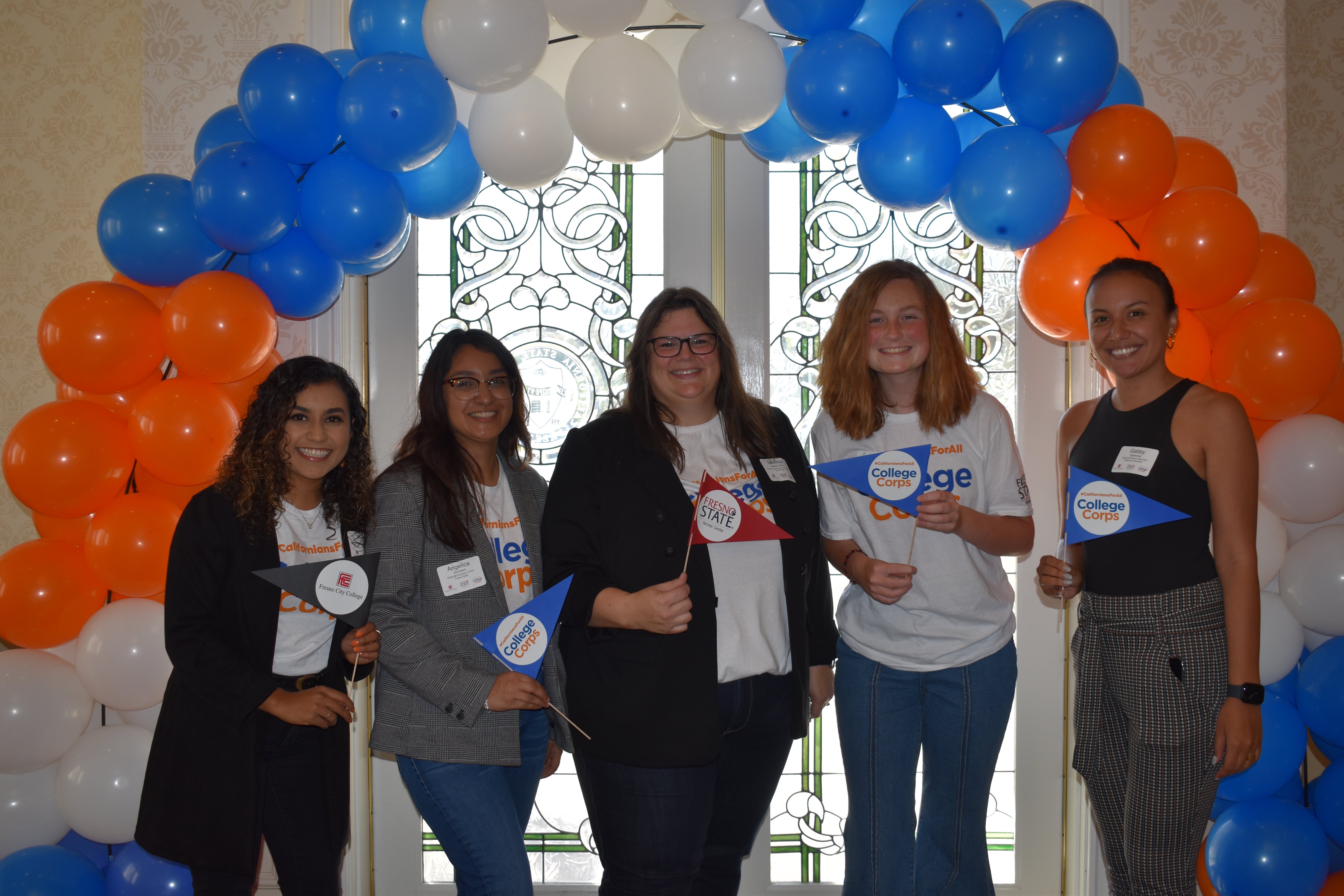 5 individuals stand abreast, smiling under a baloon arch. They are waving pennants that read College Corps, Fresno City College, or Fresno State.