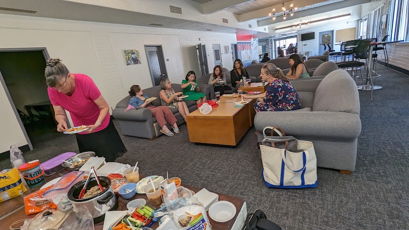 8 participants sitting on couches in the background as 1 participant looks over the various food items in the foreground