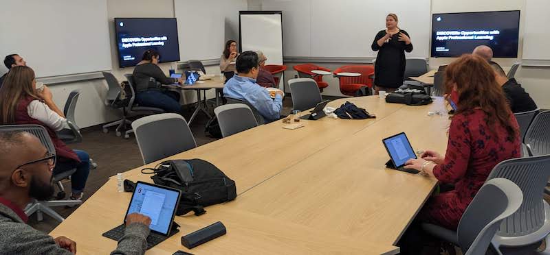 A group of 10 or so faculty working on iPads in a large meeting room as a presenter Laura Good offers instruction